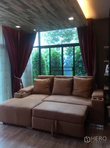 Detached House for sale in Pak Kret, Nonthaburi - Cozy lounge corner with a large brown sectional sofa beside floor-to-ceiling windows.