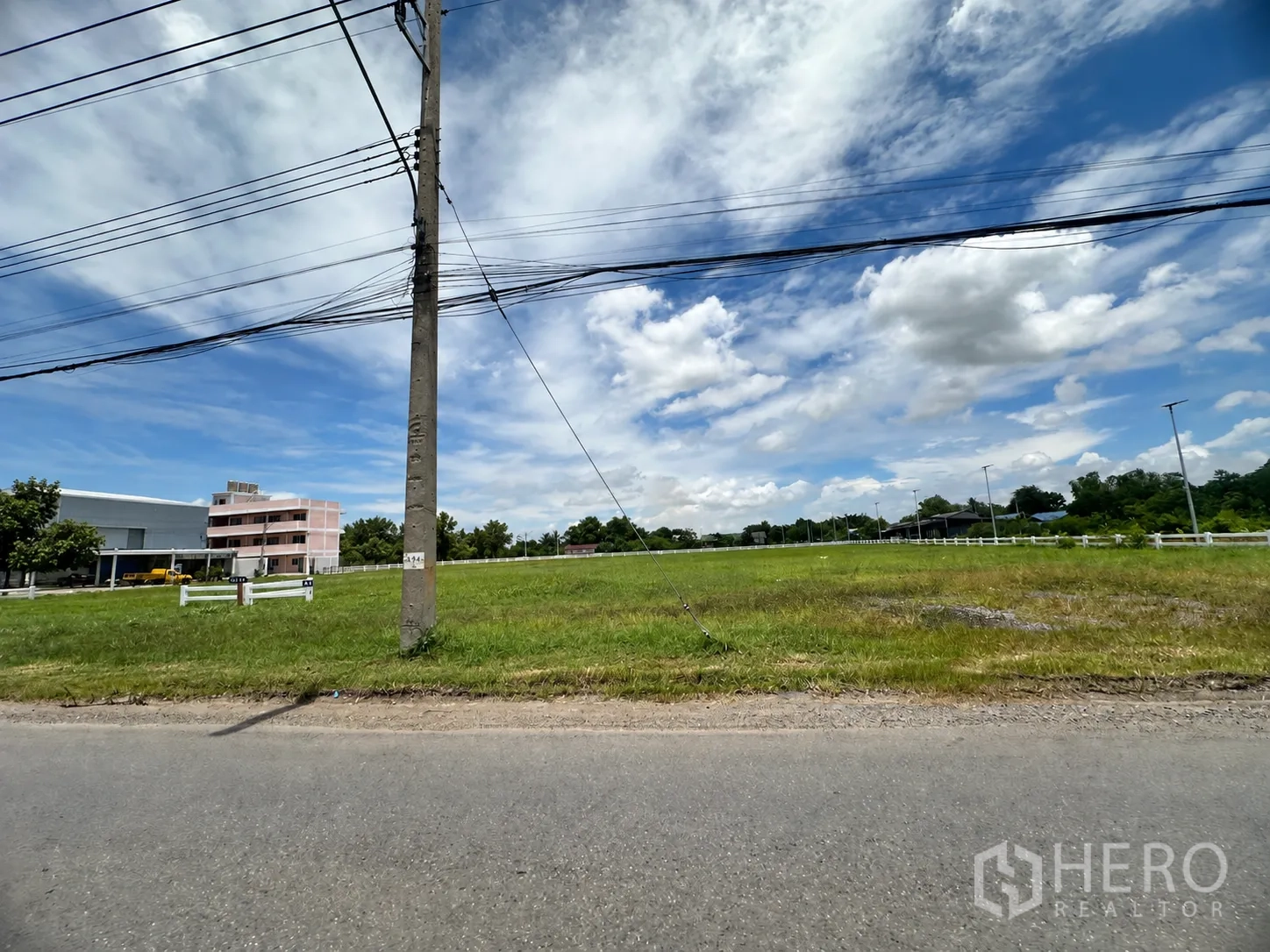 Land for sale in Lam Luk Ka, Pathum Thani - Wide-frontage grassy plot with utility pole, nearby low-rise building, and roadside fence.