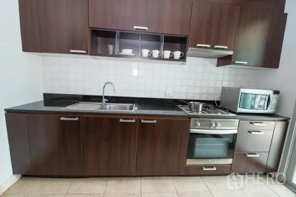 Apartment for rent in Sathon, Bangkok - Modern linear kitchen with dark wood cabinetry, built-in oven and hob, microwave, and stainless sink.