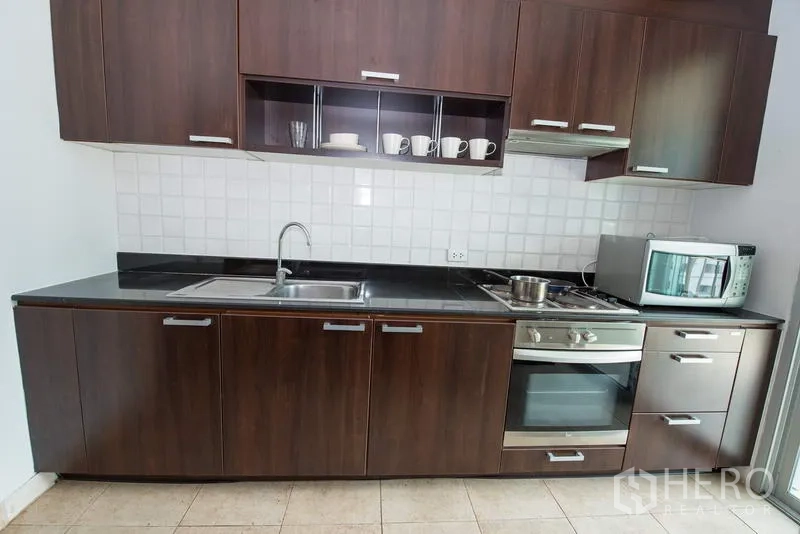 Apartment for rent in Sathon, Bangkok - Modern linear kitchen with dark wood cabinetry, built-in oven and hob, microwave, and stainless sink.