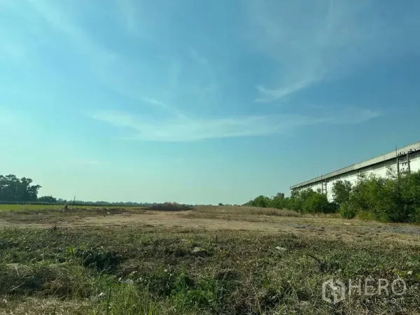 Land for sale in Chachoengsao - Expansive cleared land beside a warehouse and greenery under a bright blue sky in Chachoengsao.