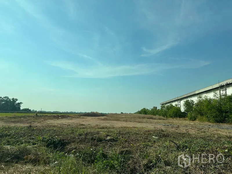 Land for sale in Chachoengsao - Expansive cleared land beside a warehouse and greenery under a bright blue sky in Chachoengsao.