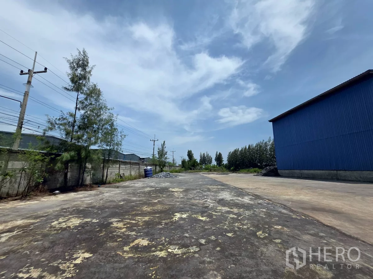 Factory for rent in Mueang Samut Sakhon, Samut Sakhon - Perimeter yard with boundary wall, power lines and trees beside a blue‑clad warehouse.