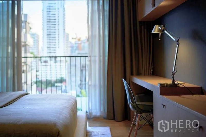 Condominium for rent in Watthana, Bangkok - Bedroom work desk facing balcony with skyline view through sheer curtains.