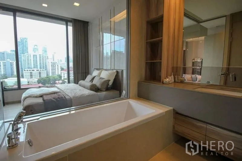 Condominium for sale in Watthana, Bangkok - Ensuite bathroom with soaking tub beside a glass window overlooking the bedroom and Bangkok city view.