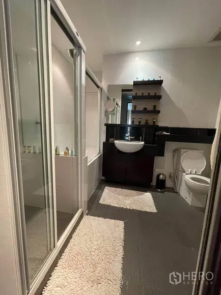 Condominium for rent or sale in Watthana, Bangkok - Bathroom with bathtub behind glass doors, vanity, open shelves and toilet.