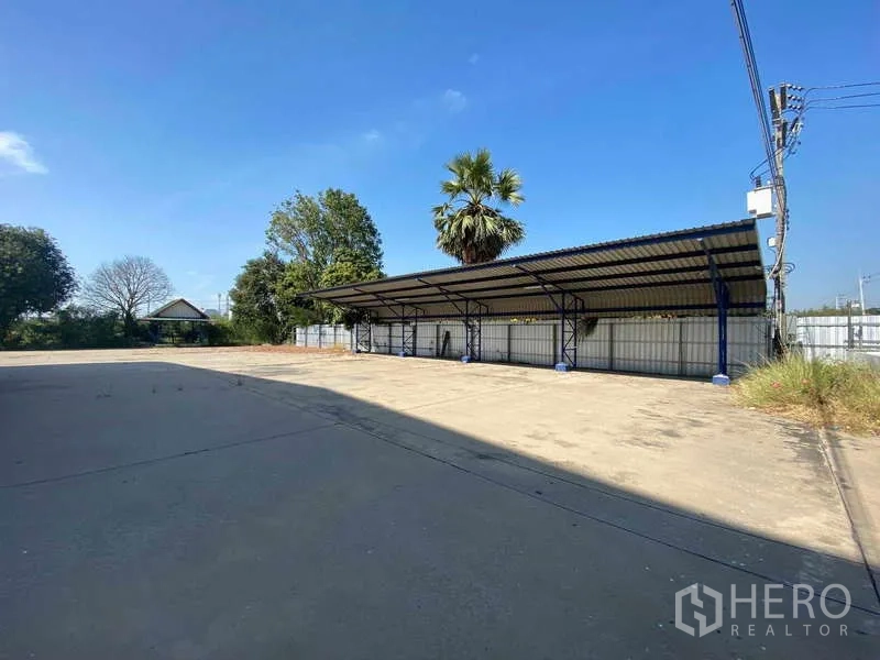 Warehouse for rent in Lat Lum Kaeo, Pathum Thani - Covered parking bays on large paved yard with fencing and trees in the background.