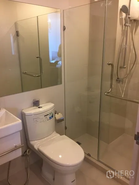 Condominium for rent in Huai Khwang, Bangkok - Modern bathroom with glass-enclosed shower, white toilet and large wall mirror.