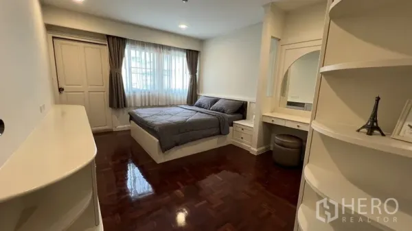 Apartment for rent in Watthana, Bangkok - Primary bedroom with large window, built-in vanity and shelving, wood floors and soft curtains.