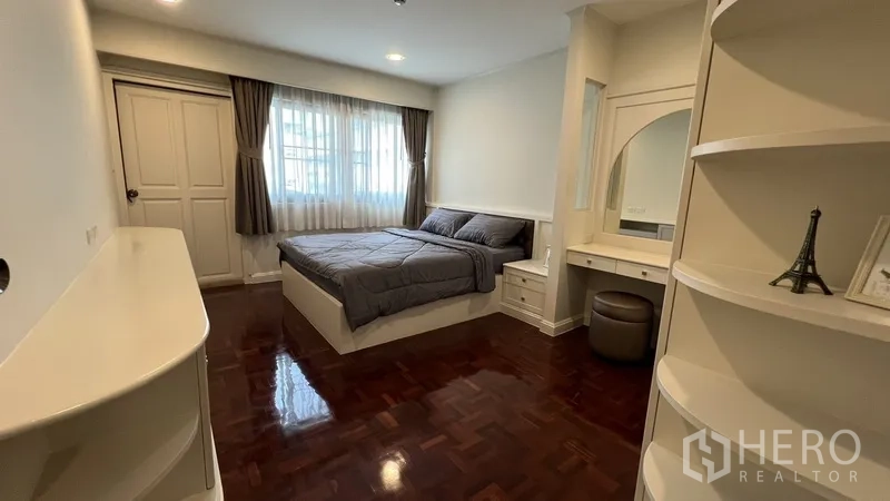 Apartment for rent in Watthana, Bangkok - Primary bedroom with large window, built-in vanity and shelving, wood floors and soft curtains.