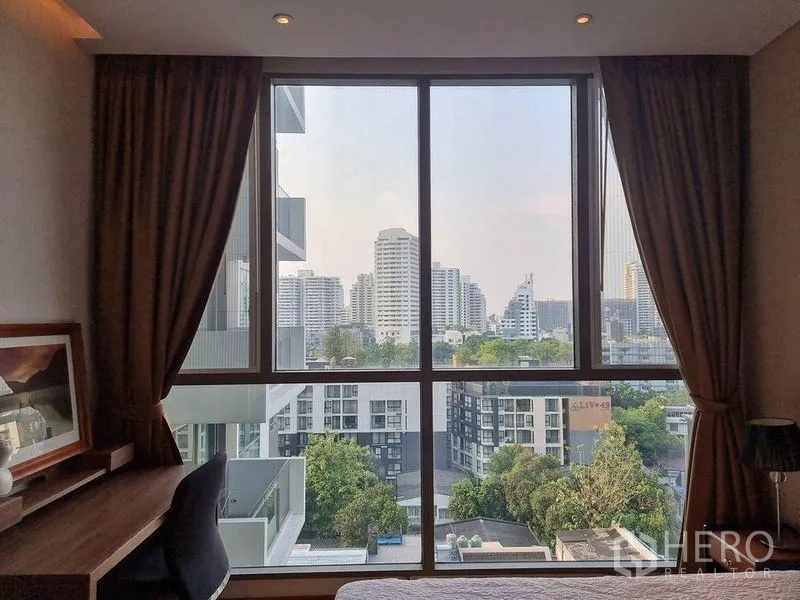 Condominium for rent in Watthana, Bangkok - City skyline view through a wide bedroom window over low-rise buildings and trees.