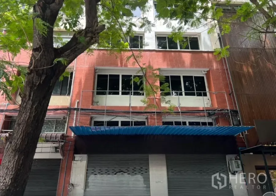 Shophouse for rent in Bang Rak, Bangkok - Four-storey shophouse facade in Bang Rak, Bangkok with red tile cladding, large windows and a blue awning beneath street trees.