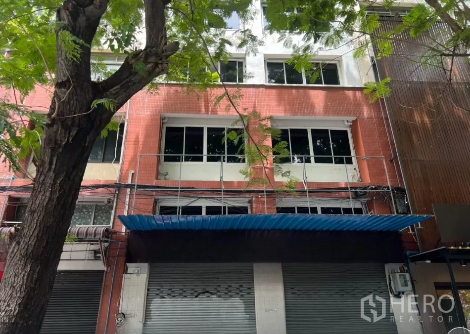 Shophouse for rent in Bang Rak, Bangkok - Four-storey shophouse facade in Bang Rak, Bangkok with red tile cladding, large windows and a blue awning beneath street trees.