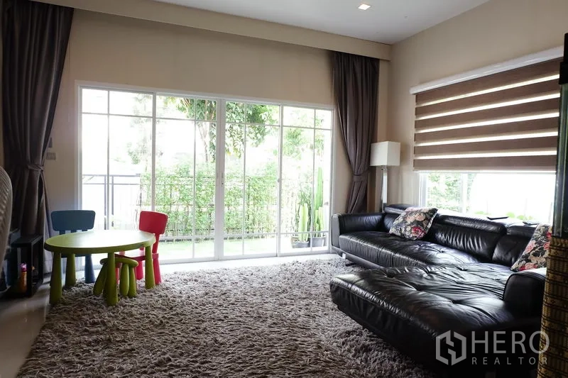 บ้าน ขายในMin Buri, Bangkok - ห้องนั่งเล่นกว้าง หน้าต่างสูงจรดเพดาน โซฟาหนัง พรมขนฟู และมู่ลี่