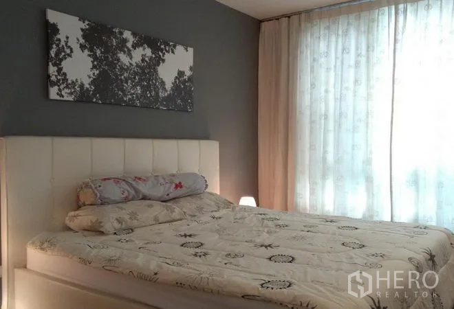 คอนโด ให้เช่าในKhlong Toei, Bangkok - ห้องนอนหลักมีหัวเตียงบุนวมสีขาว ผนังเทา และหน้าต่างบานใหญ่พร้อมม่านโปร่ง