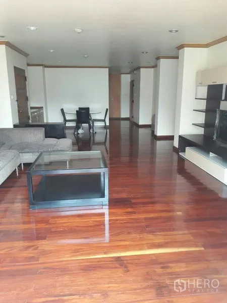 Condominium for rent in Pathum Wan, Bangkok - Spacious living and dining area with polished hardwood floors, sectional sofa, glass coffee table, and built‑in shelving.