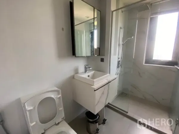 Condominium for rent in Phra Khanong, Bangkok - Contemporary bathroom with vanity, mirrored cabinet and glass-enclosed shower lit by a window.