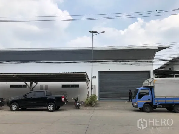 工厂 出租在Nonthaburi - 白色工厂外观，配有大型卷帘门、带顶停车位与前方送货车辆。