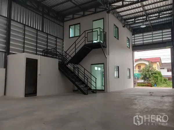 Warehouse for rent or sale in Lam Luk Ka, Pathum Thani - Warehouse interior featuring a two-storey glass-windowed office and black metal staircase, rear roller door open.