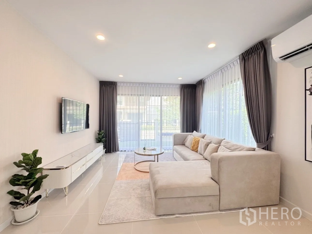 บ้าน ให้เช่าหรือขายในBang Kapi, Bangkok - ห้องนั่งเล่นผนังกระจกขนาดใหญ่ ผ้าม่านโทนเทา ทีวีติดผนัง และโซฟาตัวแอล