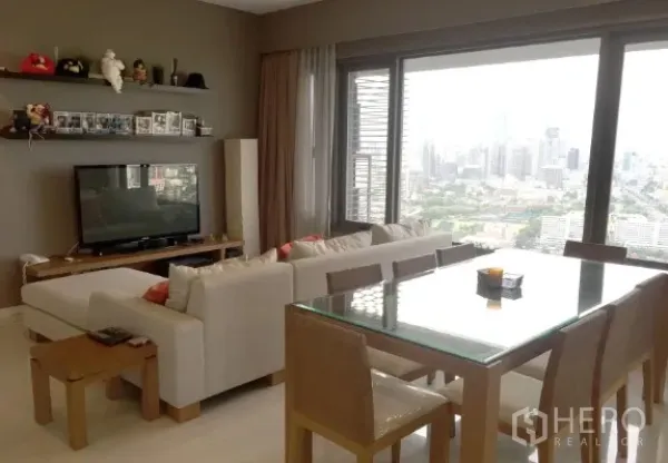 Condominium for rent or sale in Sathon, Bangkok - Living area with TV shelf and glass dining table beside floor-to-ceiling windows facing skyline and green park views.
