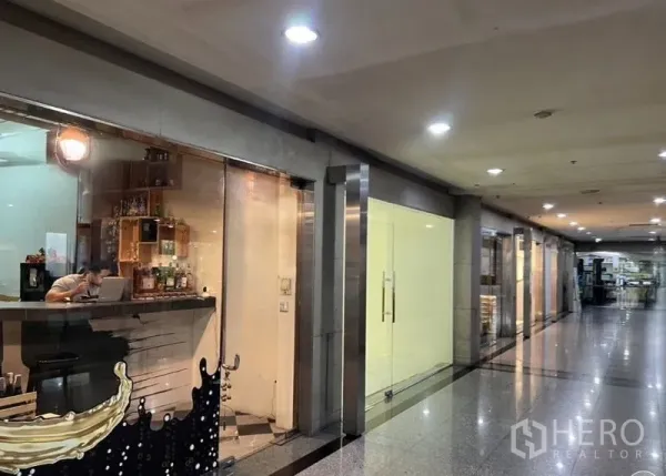 零售空间 出租在Watthana, Bangkok - 商业楼内走廊，灯光明亮的玻璃店面，旁边为待租铺位。