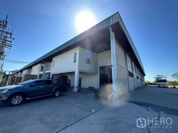 Factory for rent in Si Racha, Chon Buri - Long warehouse block with canopy, drive-through access and trucks at the loading area under bright sun.