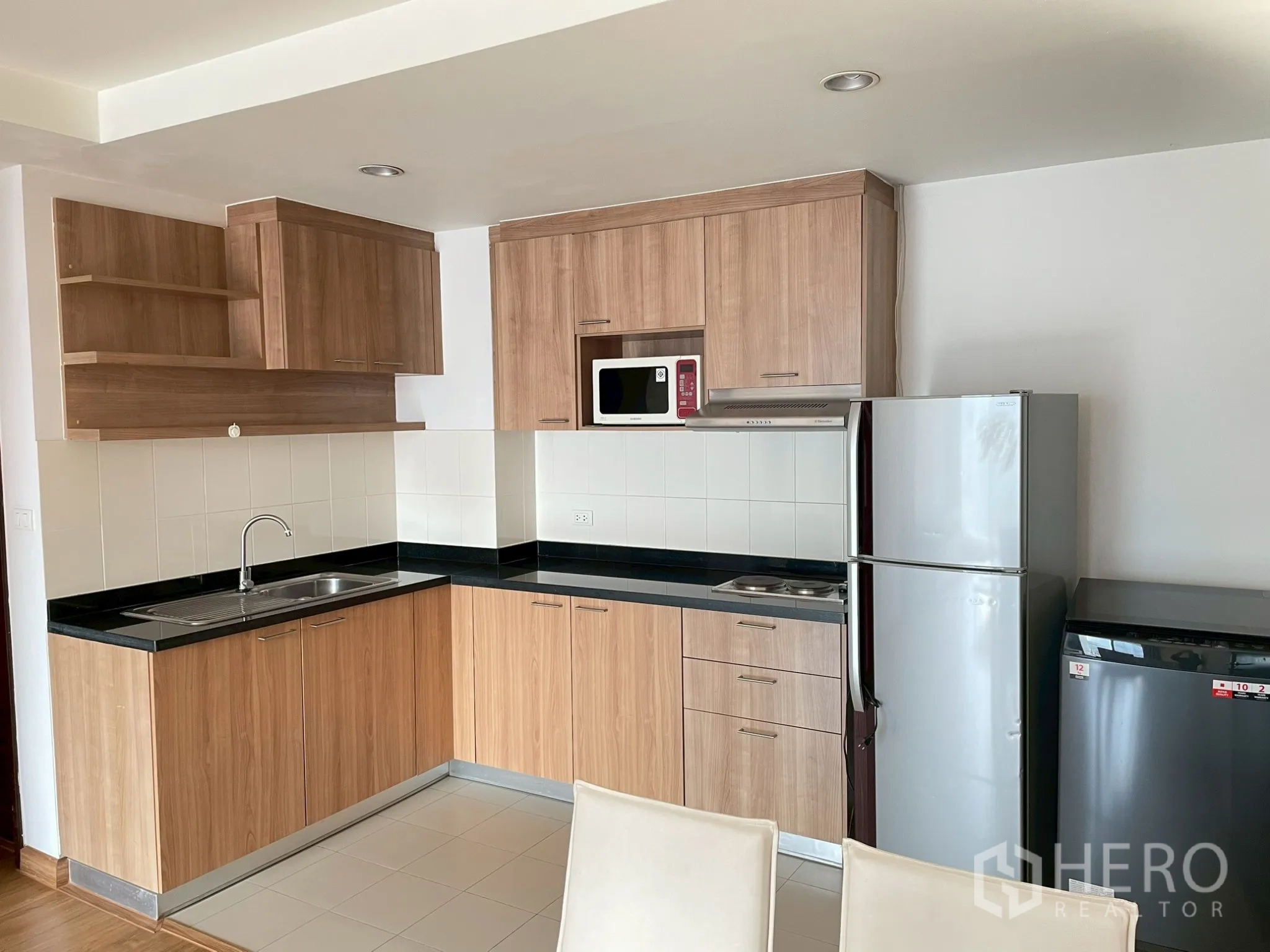 Apartment for rent in Khlong Toei, Bangkok - L-shaped kitchen with tiled backsplash, black countertops, and full-size fridge.