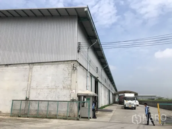货栈 出租在Bang Sao Thong, Samut Prakan - 金属外墙高挑仓库外观，宽阔混凝土场地，装卸月台与门卫岗亭，卡车出入便捷的工业区环境。