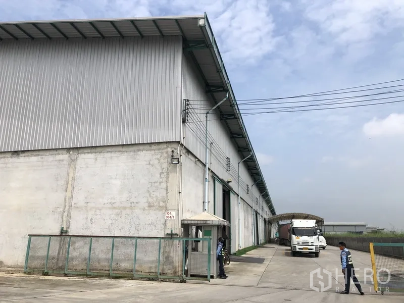 货栈 出租在Bang Sao Thong, Samut Prakan - 金属外墙高挑仓库外观，宽阔混凝土场地，装卸月台与门卫岗亭，卡车出入便捷的工业区环境。