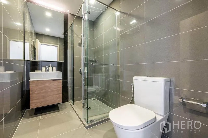 公寓 出租或出售在Watthana, Bangkok - 简约浴室，玻璃淋浴间、灰色瓷砖、洗手台与马桶。