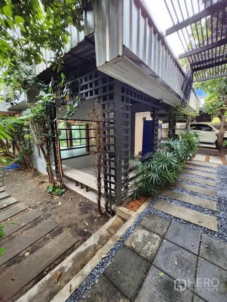 带办公室的家 出租在Lat Phrao, Bangkok - 工业风外立面一角，格栅立柱、绿化与石板路相映成趣。