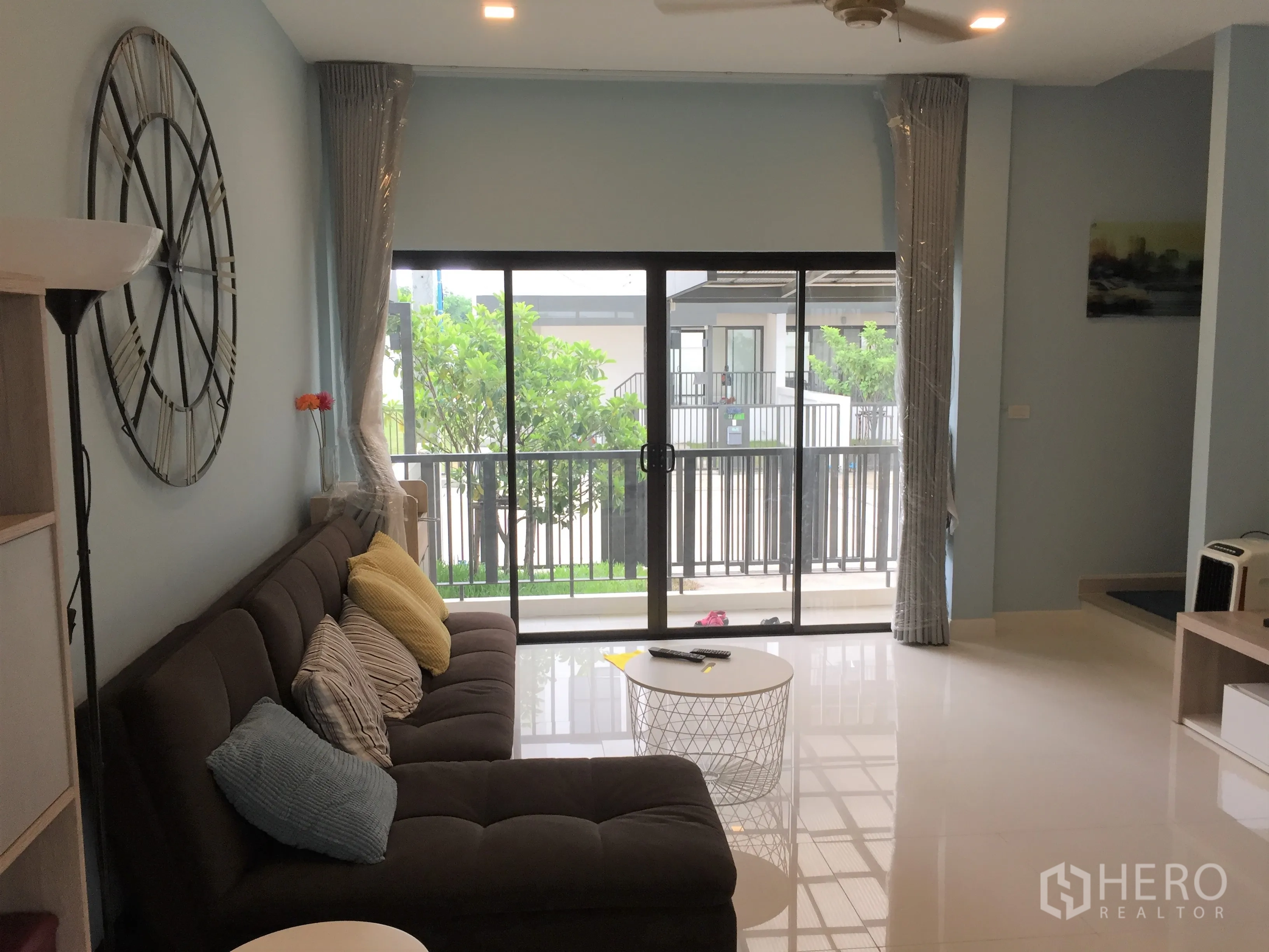 Townhouse for rent in Saphan Sung, Bangkok - Bright living room with large wall clock, sectional sofa and balcony overlooking greenery.