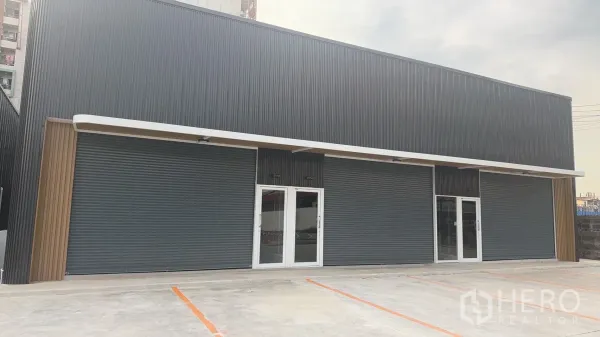 คลังสินค้า ให้เช่าในPrawet, Bangkok - ด้านหน้าโกดังสมัยใหม่ กรุลอนสีเข้ม ประตูกระจก ประตูม้วน และที่จอดรถตีเส้นชัดเจน.
