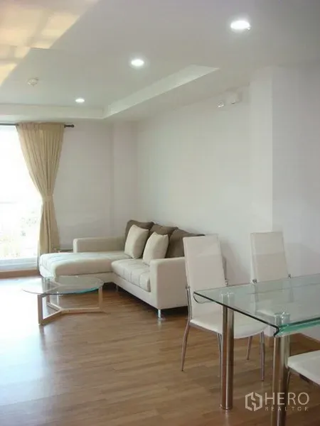 Apartment for rent in Khlong Toei, Bangkok - Bright lounge with L-shaped sofa, glass coffee table and a dining area beside a large window.