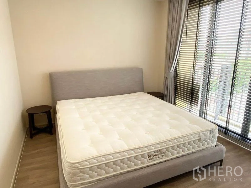 คอนโด ให้เช่าในPathum Wan, Bangkok - ห้องนอนสว่างพร้อมเตียงควีน โต๊ะข้างเตียง และหน้าต่างเต็มบานมีม่านและมู่ลี่