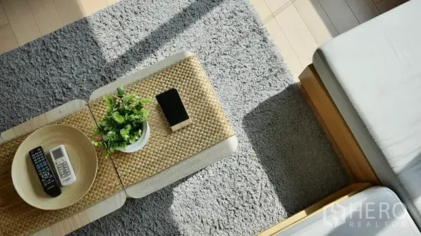 Condominium for rent or sale in Khlong Toei, Bangkok - Close-up of a woven coffee table with plant, remote and phone on a soft grey rug between sofas.