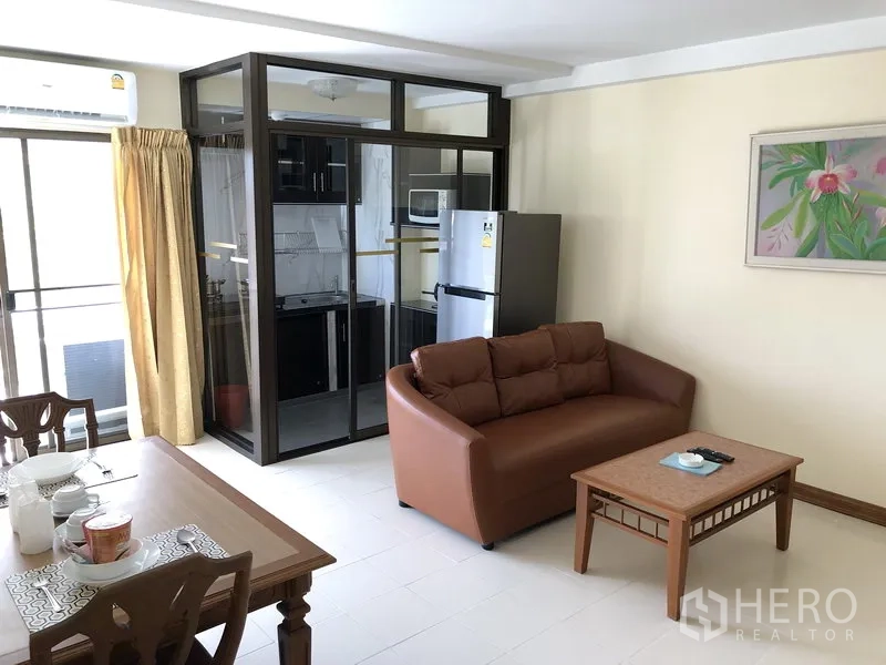 Apartment for rent in Suan Luang, Bangkok - Open-plan lounge with a glass-enclosed kitchen, refrigerator and dining table beside balcony doors.