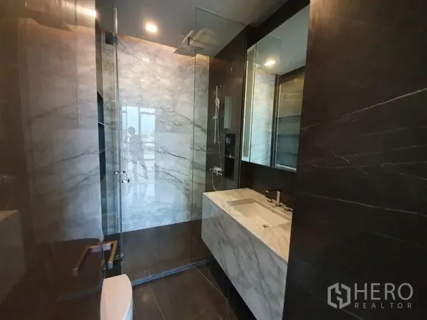 Condominium for rent in Watthana, Bangkok - Modern bathroom with glass-enclosed rain shower, marble vanity and dark stone walls.