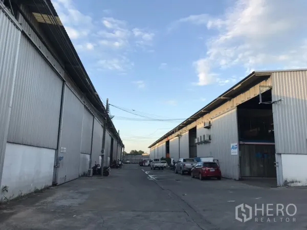 Warehouse for rent in Phra Pradaeng, Samut Prakan - Row of corrugated metal warehouses with a wide driveway and parked vehicles under a blue sky.