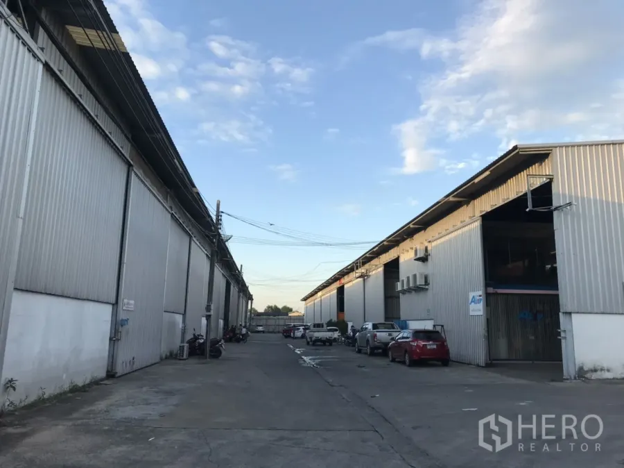 Warehouse for rent in Phra Pradaeng, Samut Prakan - Row of corrugated metal warehouses with a wide driveway and parked vehicles under a blue sky.