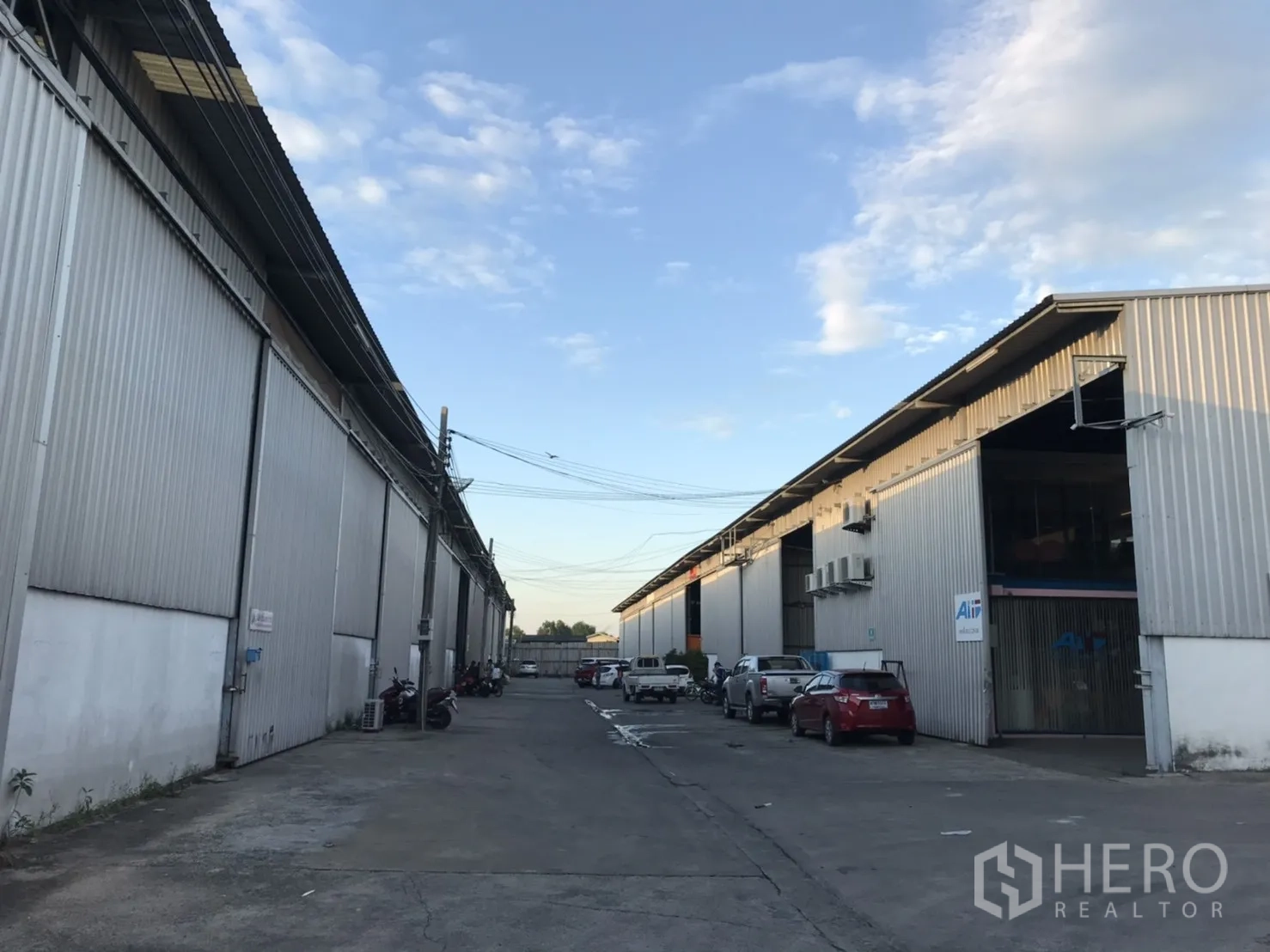 Warehouse for rent in Phra Pradaeng, Samut Prakan - Row of corrugated metal warehouses with a wide driveway and parked vehicles under a blue sky.