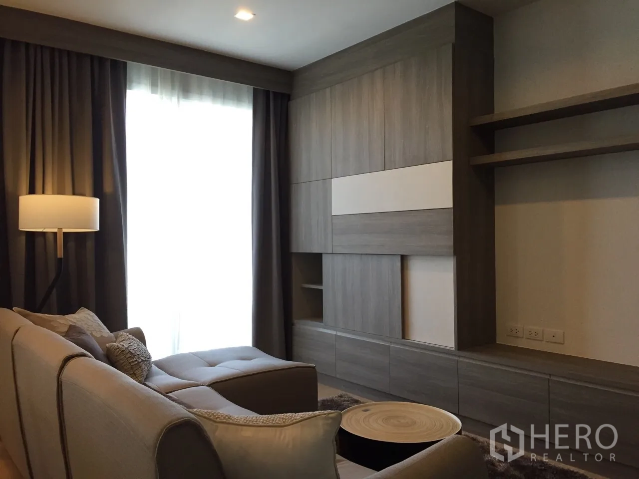 Condominium for rent or sale in Watthana, Bangkok - Modern living room with L‑shaped sofa, built‑in media wall, floor lamp and large curtained window.