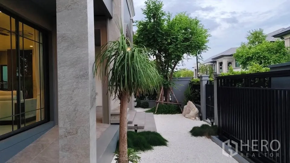 Detached House for rent or sale in Lat Krabang, Bangkok - Minimalist side garden with gravel, lush plants, and a black metal gate.