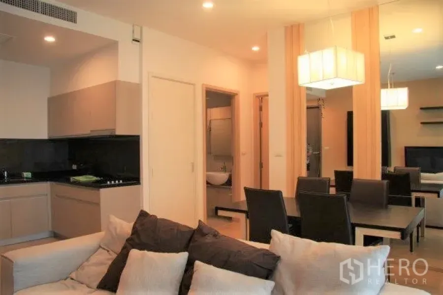 Condominium for rent in Watthana, Bangkok - Open-plan living, dining and kitchen with beige cabinetry, black backsplash and pendant lighting leading to the bedrooms and bathroom.
