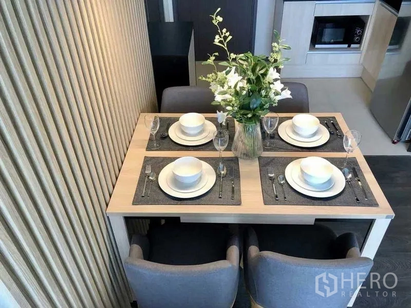 Condominium for rent in Watthana, Bangkok - Overhead view of a dining table set for four with placemats and flowers next to ribbed wall panels and kitchen.