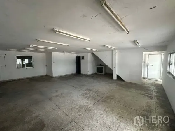 工厂 出租在Nonthaburi - 一层空置办公室，荧光灯照明，水泥地面并带楼梯通往二层。