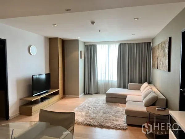 公寓 出租在Watthana, Bangkok - 温馨起居区，配转角沙发、长绒地毯、电视柜与落地窗帘。