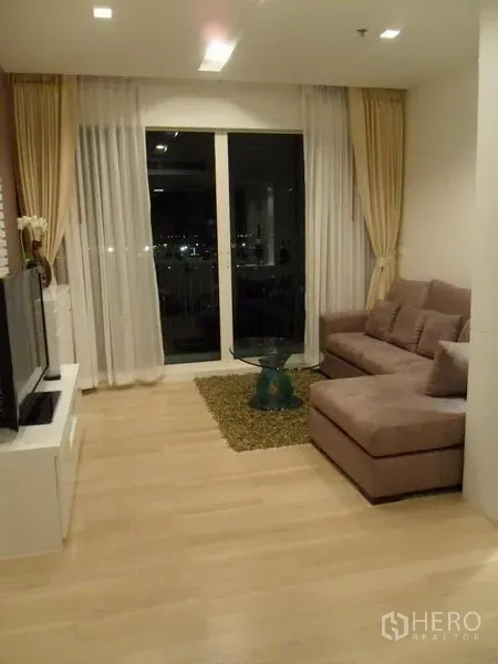 คอนโด ให้เช่าในKhlong Toei, Bangkok - ห้องนั่งเล่นโซฟา L‑shape โต๊ะกลางกระจก และประตูกระจกออกระเบียงเห็นวิวเมืองยามค่ำ