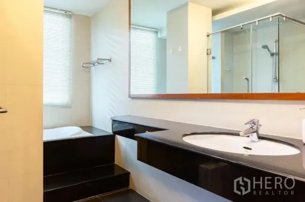 Condominium for rent in Bang Rak, Bangkok - Bathroom with long vanity, large mirror, step-up bathtub by the window and adjacent glass shower area.